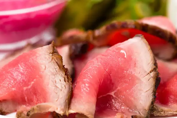 Fette sottili di roast beef ricetta classica, succulente e rosate, con un bordo croccante. Sullo sfondo, un contorno rosa e verde sfocato.
